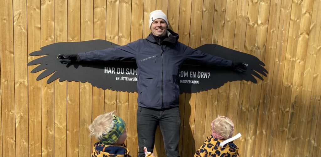 man standing in front of two kids stretching his arms wide. behind him is a wall with the silhouette of of a large bird